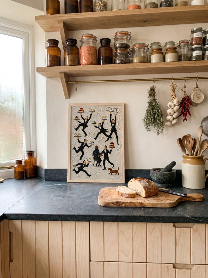 May include: A rectangular framed wall art piece featuring a textured off-white background with multiple illustrated figures in black tuxedos carrying trays of food and drinks, a seated person at a small table, and a small brown dog at the bottom.