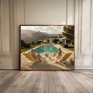 May include: A rectangular wall art print in a thin brown frame depicting a sunny outdoor scene with two people lounging on yellow chaise chairs by a turquoise swimming pool, with a modern house, green trees, and distant mountains in the background.