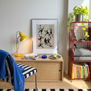 May include: A framed art print featuring a whimsical illustration of multiple waiters in black suits carrying trays of food and drinks, with a small dog in the corner, displayed in a silver frame on a light wood cabinet next to a yellow desk lamp.