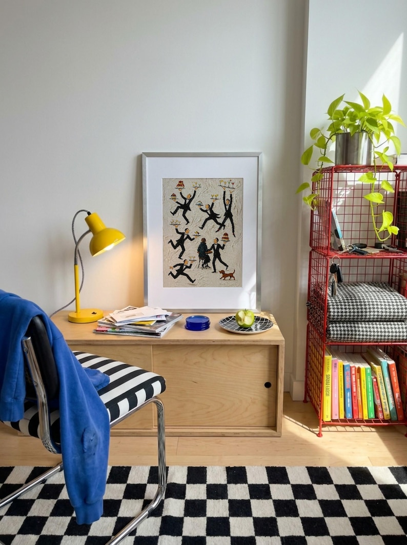 May include: A framed art print featuring a whimsical illustration of multiple waiters in black suits carrying trays of food and drinks, with a small dog in the corner, displayed in a silver frame on a light wood cabinet next to a yellow desk lamp.