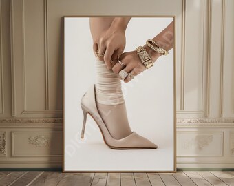 May include: A framed photograph featuring a high-heeled shoe, sheer sock, and jewelry. The shoe is a light beige color with a stiletto heel. The hands are adorned with rings and bracelets. The background is a neutral color.