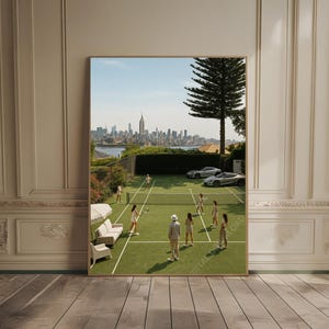 May include: A framed print depicts a tennis court scene with players in white attire. The green court is set against a backdrop of a city skyline and a clear blue sky. Luxury cars are parked nearby. A sofa and chair are on the side of the court.