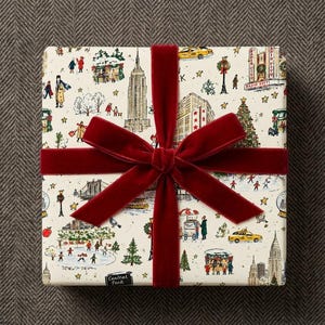 May include: A square gift box wrapped in cream-colored paper featuring a colorful New York City illustration with landmarks like the Empire State Building and Radio City, tied with a wide, deep red velvet ribbon and a large bow.