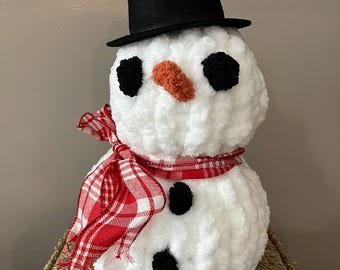 May include: A plush snowman crafted from white yarn, featuring a black felt top hat, two black pom-pom eyes, an orange textured nose, and two black pom-pom buttons on the body. A red and white plaid fabric scarf is tied around the neck.