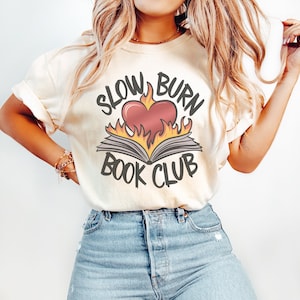 May include: A white t-shirt with the text "Slow Burn Book Club" in black lettering. A red heart with flames is on top of an open book.