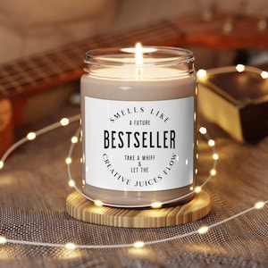 May include: A brown candle with a white label that reads "Smells Like A Future Bestseller Take A Whiff & Let The Creative Juices Flow". The candle is lit and sitting on a wooden coaster.