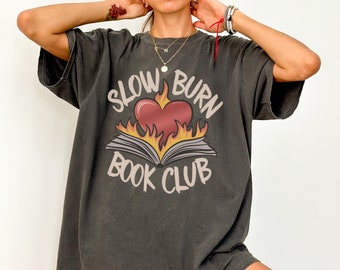 Puede incluir: Una camiseta de gran tamaño de color gris oscuro con un estampado gráfico de un corazón en llamas con un libro debajo. El texto "Slow Burn Book Club" está impreso alrededor del gráfico.