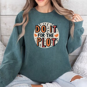 May include: Teal blue sweatshirt with a white circle graphic that says "Do It For The Plot" in orange and black lettering with two small red hearts.