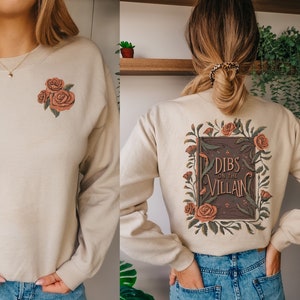 May include: A beige sweatshirt with a floral design on the front and back. The front design features three pink roses. The back design features a book with the text "Dibs on the Villain" surrounded by flowers.