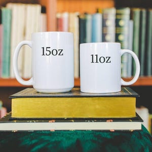 Puede incluir: Dos tazas de cer&aacute;mica blancas, etiquetadas como "15oz" y "11oz", est&aacute;n sobre una pila de libros. Las tazas tienen asas. El fondo presenta una estanter&iacute;a llena de libros.