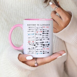 Puede incluir: Una taza de cer&aacute;mica blanca con asa y borde rosas. La taza presenta un dise&ntilde;o de tarjeta de biblioteca vintage con el texto "RETURN TO LIBRARY" y varias fechas. La taza est&aacute; sostenida por una persona.