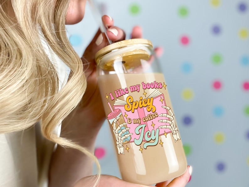 Bookish iced coffee cup with bamboo lid and straw. Groovy retro text in pastel colors reads, “I like my books spicy & my coffee icy.” Skeleton hands hold an iced coffee mug behind an open book.