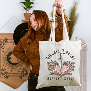 May include: A white canvas tote bag with the text "Villain Lovers Support Group" printed in black. A black and white illustration of a book with a dagger sticking out of it and roses on either side is above the text.