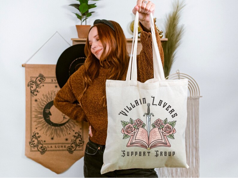 May include: A white canvas tote bag with the text "Villain Lovers Support Group" printed in black. A black and white illustration of a book with a dagger sticking out of it and roses on either side is above the text.