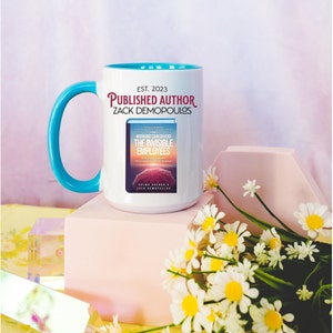 May include: White ceramic mug with a turquoise handle. The mug has the text "EST. 2023 PUBLISHED AUTHOR ZACK DEMOPOULOS" printed on it. The mug is sitting on a pink surface with a bouquet of white daisies in the foreground.