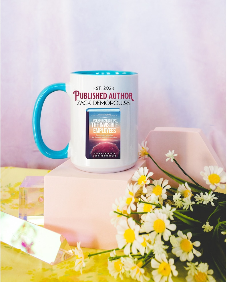 May include: White ceramic mug with a turquoise handle. The mug has the text "EST. 2023 PUBLISHED AUTHOR ZACK DEMOPOULOS" printed on it. The mug is sitting on a pink surface with a bouquet of white daisies in the foreground.