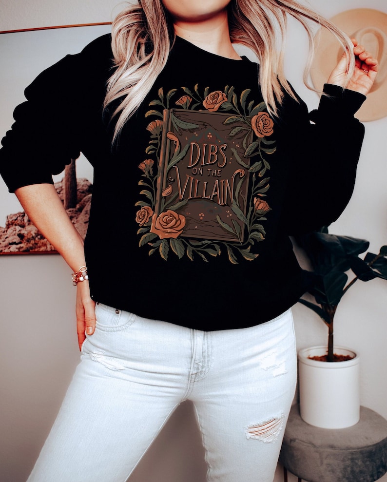 Book merch crewneck sweatshirt with a graphic of a book and dark florals and “dibs on the villain” text