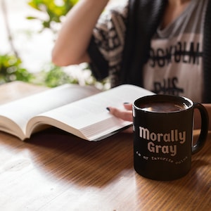 Pu&ograve; includere: Una tazza da caff&egrave; nera con la scritta "Morally Gray is my favorite color". La tazza &egrave; appoggiata su un tavolo di legno accanto a un libro aperto.
