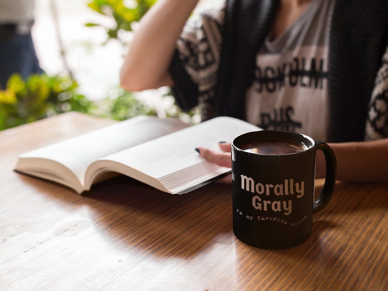 Pu&ograve; includere: Una tazza da caff&egrave; nera con la scritta "Morally Gray is my favorite color". La tazza &egrave; appoggiata su un tavolo di legno accanto a un libro aperto.