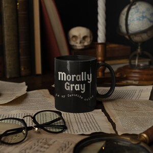 Pu&ograve; includere: Una tazza da caff&egrave; nera con la scritta "Morally Gray is my favorite color". La tazza &egrave; appoggiata su un tavolo con una lente d'ingrandimento, un teschio, un mappamondo e una candela accesa.