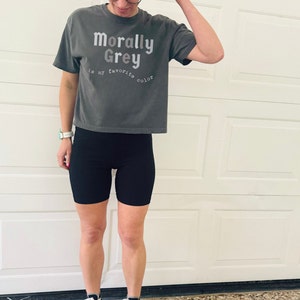 May include: A woman wearing a gray cropped t-shirt with the text "Morally Grey is my favorite color" in white. She is also wearing black biker shorts and black and white high-top sneakers.