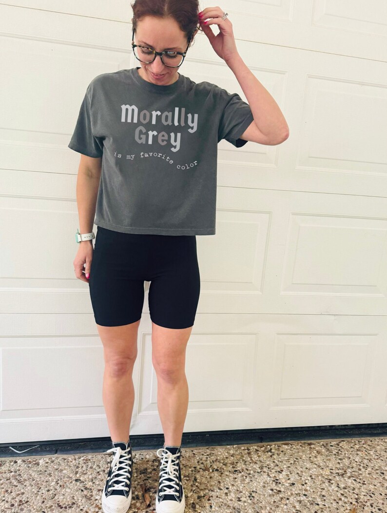 May include: A woman wearing a gray cropped t-shirt with the text "Morally Grey is my favorite color" in white. She is also wearing black biker shorts and black and white high-top sneakers.