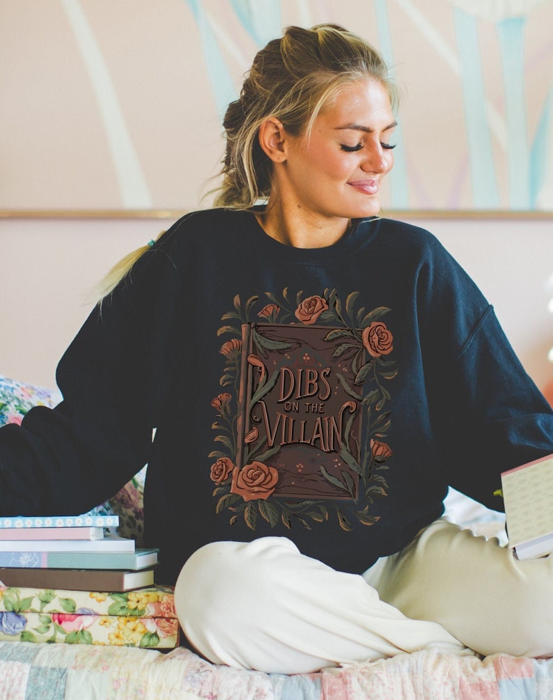 Book merch crewneck sweatshirt with a graphic of a book and dark florals and “dibs on the villain” text
