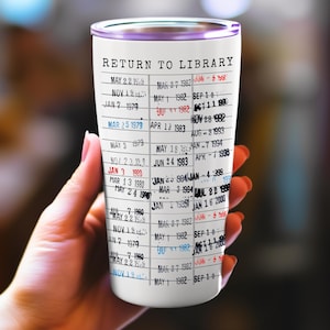 May include: A white tumbler with a purple rim, featuring a vintage library card design. The text "RETURN TO LIBRARY" is at the top, with rows of dates and numbers resembling a card catalog. The tumbler is held by a hand.