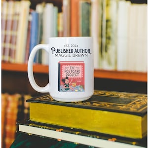 May include: White ceramic mug with a black handle. The mug has the text "EST. 2024 PUBLISHED AUTHOR MAGGIE BROWN" and an illustration of a book with the text "THE POSTCARD PROJECT".