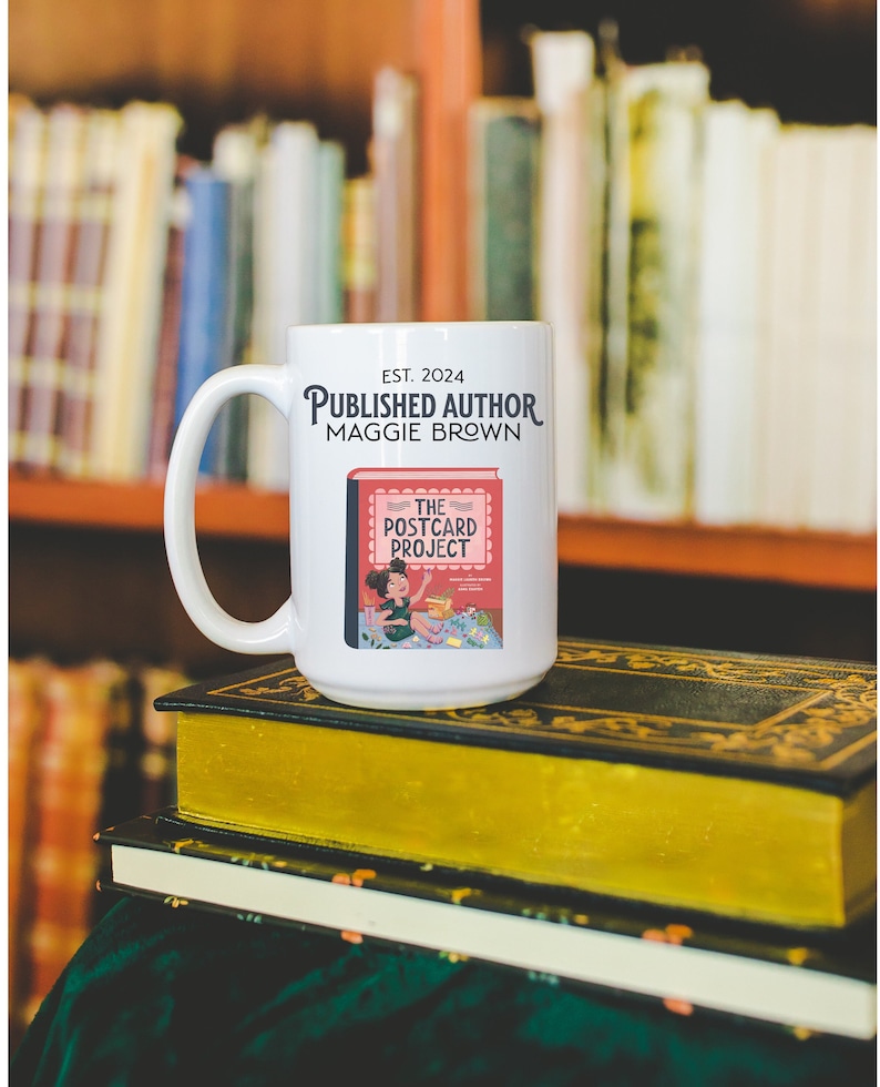 May include: White ceramic mug with a black handle. The mug has the text "EST. 2024 PUBLISHED AUTHOR MAGGIE BROWN" and an illustration of a book with the text "THE POSTCARD PROJECT".