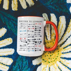 Puede incluir: Taza de cer&aacute;mica blanca con asa y borde rojos, con un dise&ntilde;o de "Return to Library" con fechas y texto. La taza se encuentra sobre un fondo con estampado floral colorido.