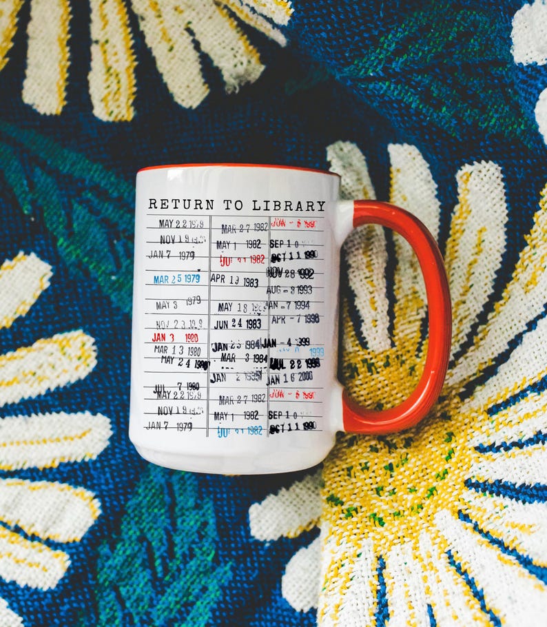 Puede incluir: Taza de cer&aacute;mica blanca con asa y borde rojos, con un dise&ntilde;o de "Return to Library" con fechas y texto. La taza se encuentra sobre un fondo con estampado floral colorido.