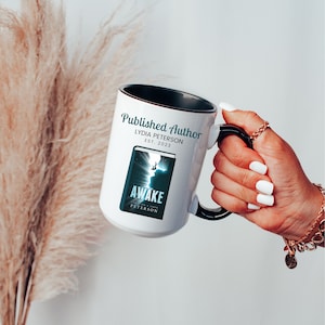 May include: White ceramic mug with a black handle and rim, featuring the text "Published Author" and "Awake" with a book cover design. The mug is held by a hand with white nail polish, and a gold bracelet.