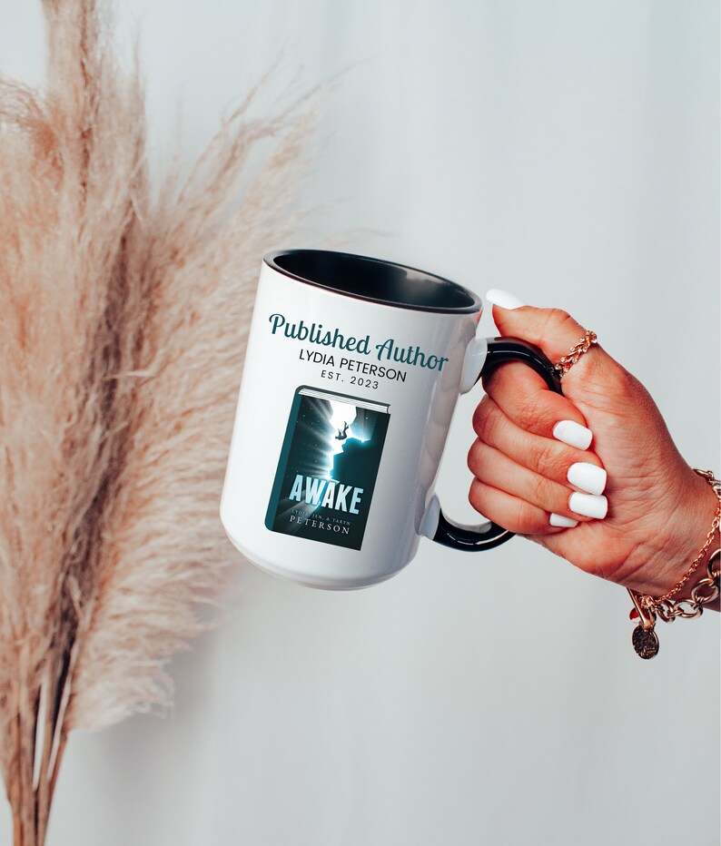 May include: White ceramic mug with a black handle and rim, featuring the text "Published Author" and "Awake" with a book cover design. The mug is held by a hand with white nail polish, and a gold bracelet.