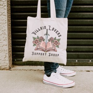 May include: A white canvas tote bag with the text "Villain Lovers Support Group" and a graphic of an open book with a sword through it and pink flowers.