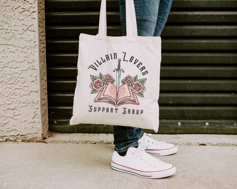 May include: A white canvas tote bag with the text "Villain Lovers Support Group" and a graphic of an open book with a sword through it and pink flowers.