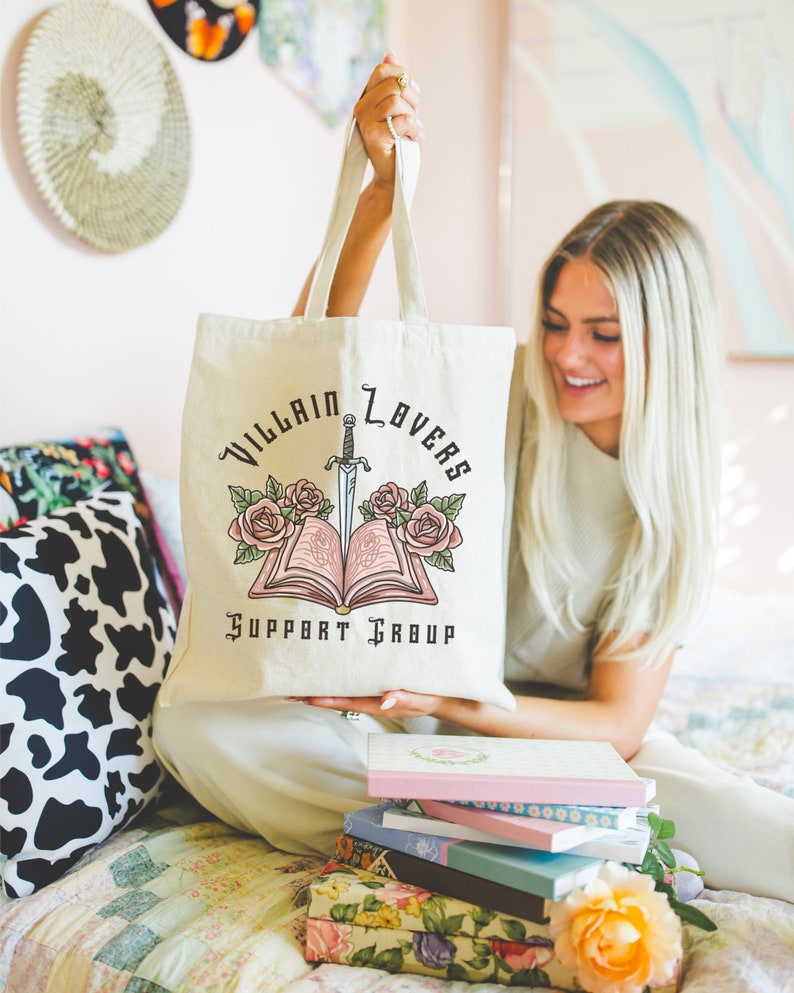 May include: A white canvas tote bag with a black and pink design. The design features a book with a dagger sticking out of it, surrounded by roses. The text on the bag reads "Villain Lovers Support Group".