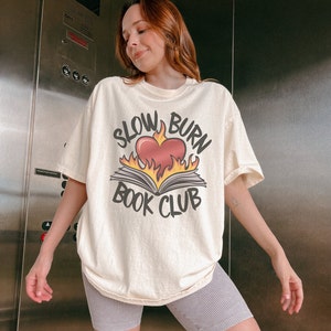 May include: A woman wearing a cream-coloured t-shirt with a graphic of a book on fire and the text "Slow Burn Book Club". She is also wearing grey striped cycling shorts.