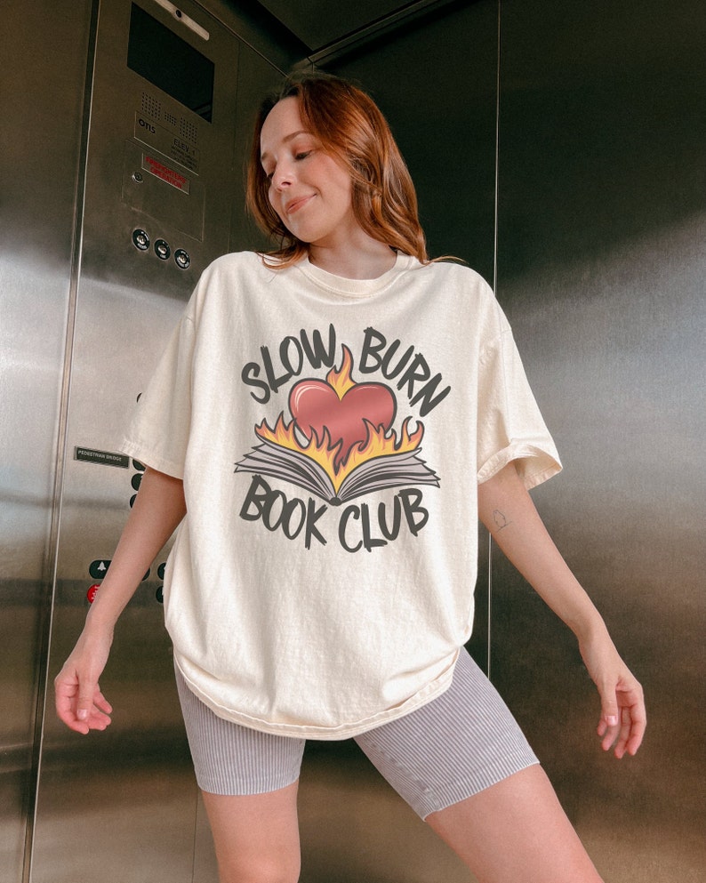 May include: A woman wearing a cream-coloured t-shirt with a graphic of a book on fire and the text "Slow Burn Book Club". She is also wearing grey striped cycling shorts.