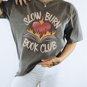 May include: A grey t-shirt with a graphic of a book on fire with a heart in the middle. The text "Slow Burn Book Club" is printed on the shirt.