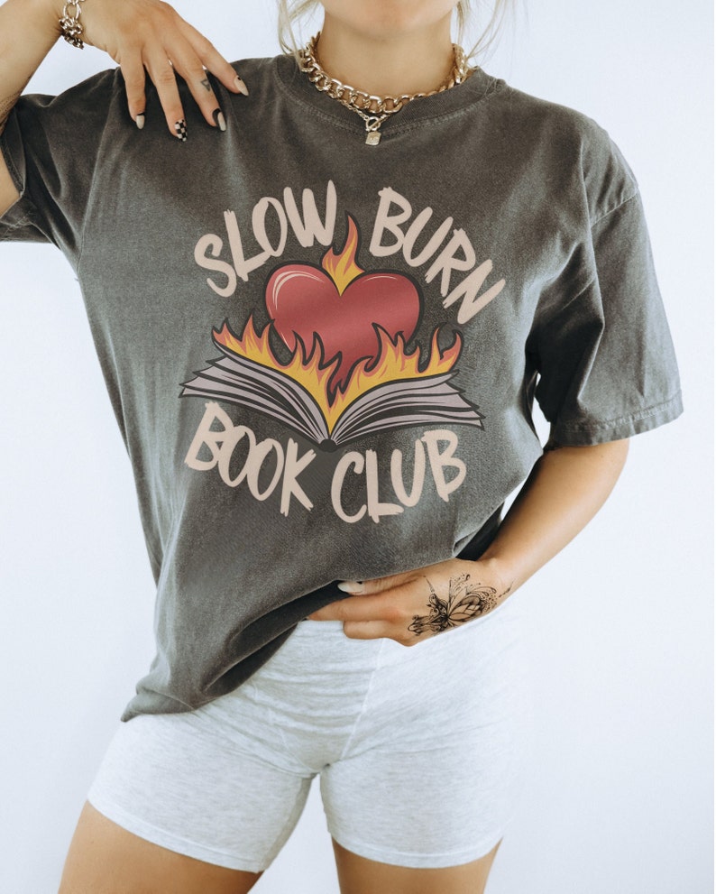 May include: A grey t-shirt with a graphic of a book on fire with a heart in the middle. The text "Slow Burn Book Club" is printed on the shirt.