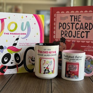 May include: A children's book and two mugs featuring the text "Joy The Pandacorn" and "The Postcard Project." The book covers are pink and white with colorful illustrations. The mugs are white with black and pink accents, and the text "Published Author" is printed on them.