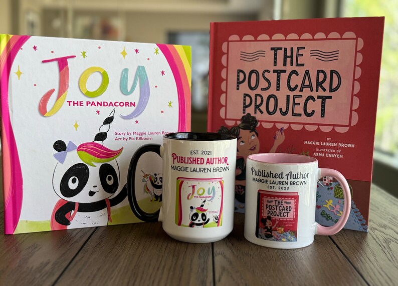 May include: A children's book and two mugs featuring the text "Joy The Pandacorn" and "The Postcard Project." The book covers are pink and white with colorful illustrations. The mugs are white with black and pink accents, and the text "Published Author" is printed on them.