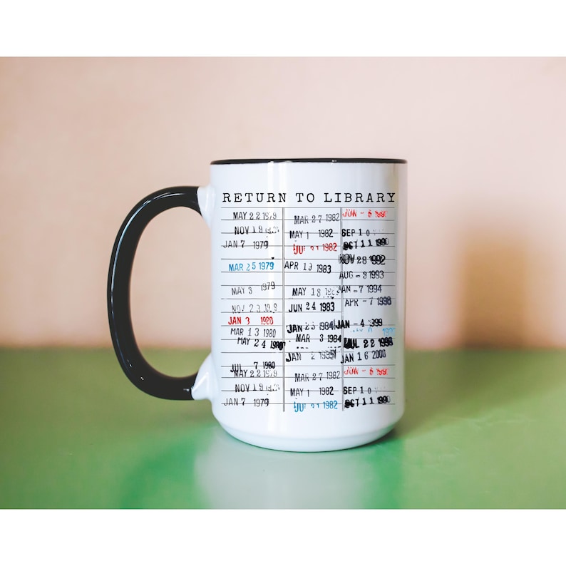 Puede incluir: Taza de cer&aacute;mica blanca con asa y borde negros. La taza presenta un dise&ntilde;o impreso que se asemeja a una tarjeta de biblioteca, con el texto "RETURN TO LIBRARY" y varias fechas en rojo, azul y negro.