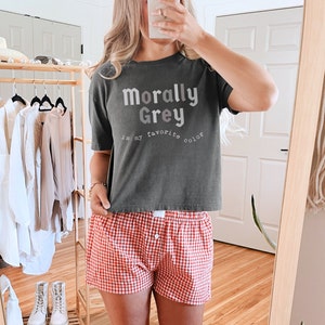 May include: A woman wearing a gray t-shirt with the text "Morally Grey is my favorite color" and red and white checkered shorts.