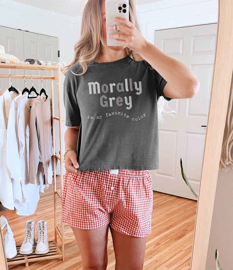 May include: A woman wearing a gray t-shirt with the text "Morally Grey is my favorite color" and red and white checkered shorts.