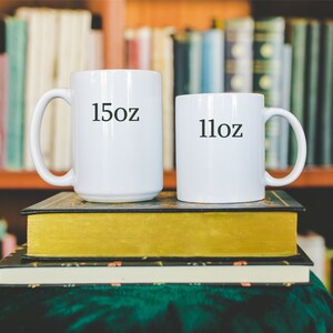 May include: Two white ceramic mugs with black text. The larger mug says "15oz" and the smaller mug says "11oz". The mugs are stacked on top of two books.