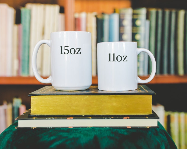 May include: Two white ceramic mugs with black text. The larger mug says "15oz" and the smaller mug says "11oz". The mugs are stacked on top of two books.