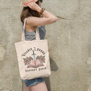 May include: A beige tote bag with a design of a book with a sword through it and roses on either side. The text on the bag reads "Villain Lovers Support Group".
