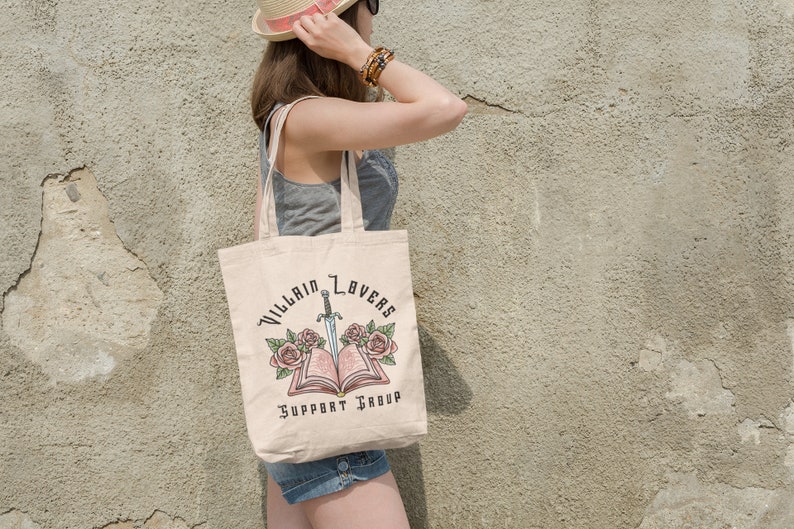 May include: A beige tote bag with a design of a book with a sword through it and roses on either side. The text on the bag reads "Villain Lovers Support Group".